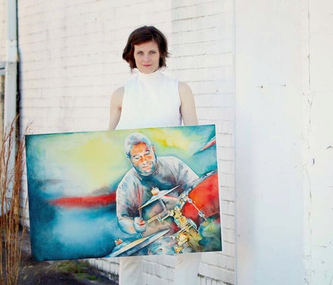 A person standing in front of a white wall, holding up a large artwork featuring a colorful illustration of a man playing a drum set.