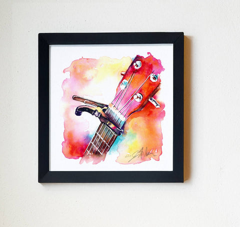 A vibrant watercolor painting of a ukulele headstock with a capo, featuring bright pink-coral hues and other colors, displayed in a white frame.