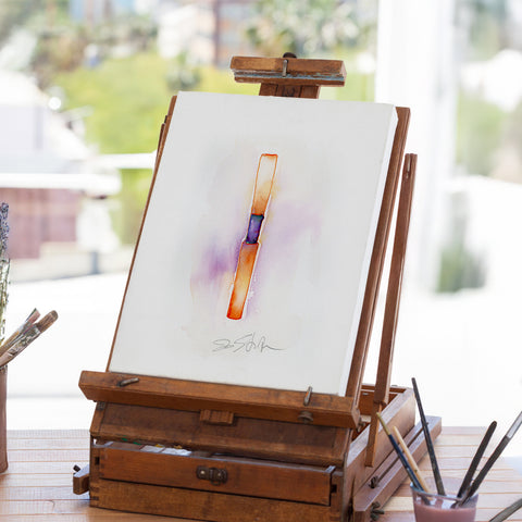 Wooden easel with a painting of a reed on a white background, set against a blurred outdoor scene.