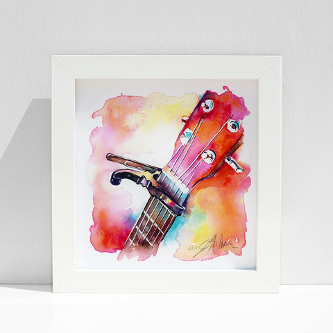 A vibrant watercolor painting of a guitar headstock with a capo, featuring bright pink-coral hues and other colors, displayed in a white frame.