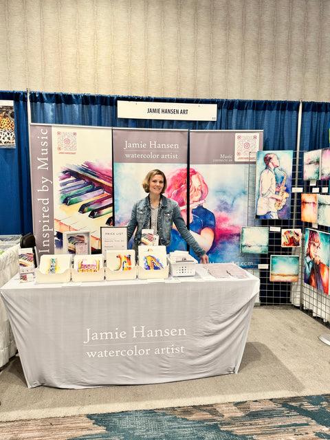 Jamie Hansen stands behind a table displaying her music-inspired watercolor artwork at a conference booth, surrounded by colorful prints and signage